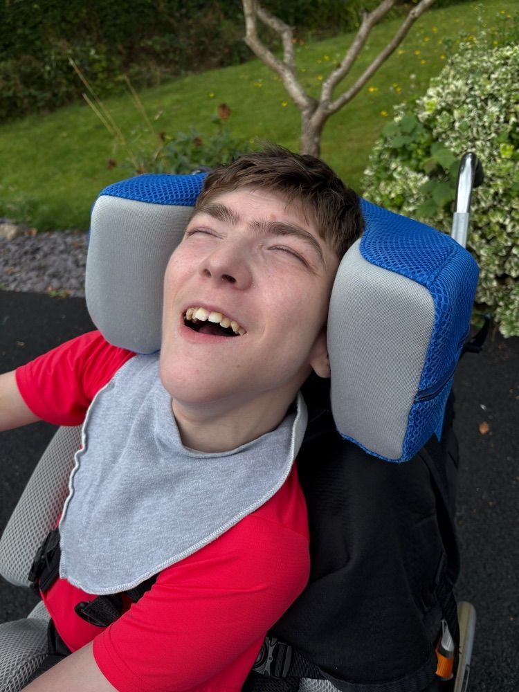 Smiling person in wheelchair, wearing bib. Red shirt, gray headrest. Outdoors with greenery.
