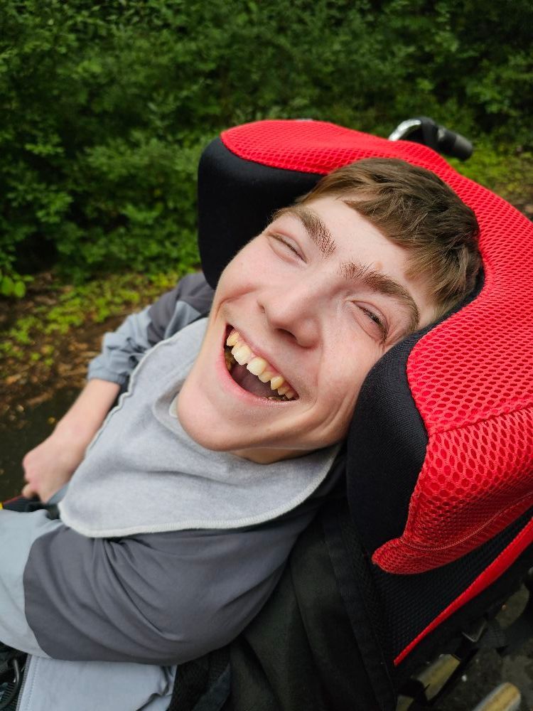 Young man with a wide smile in a wheelchair, wearing a gray bib. Red headrest, outdoors with trees.