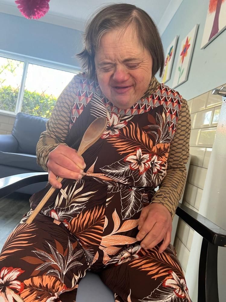 Woman with Down syndrome in floral jumpsuit, holding wooden spoon, smiling in kitchen.