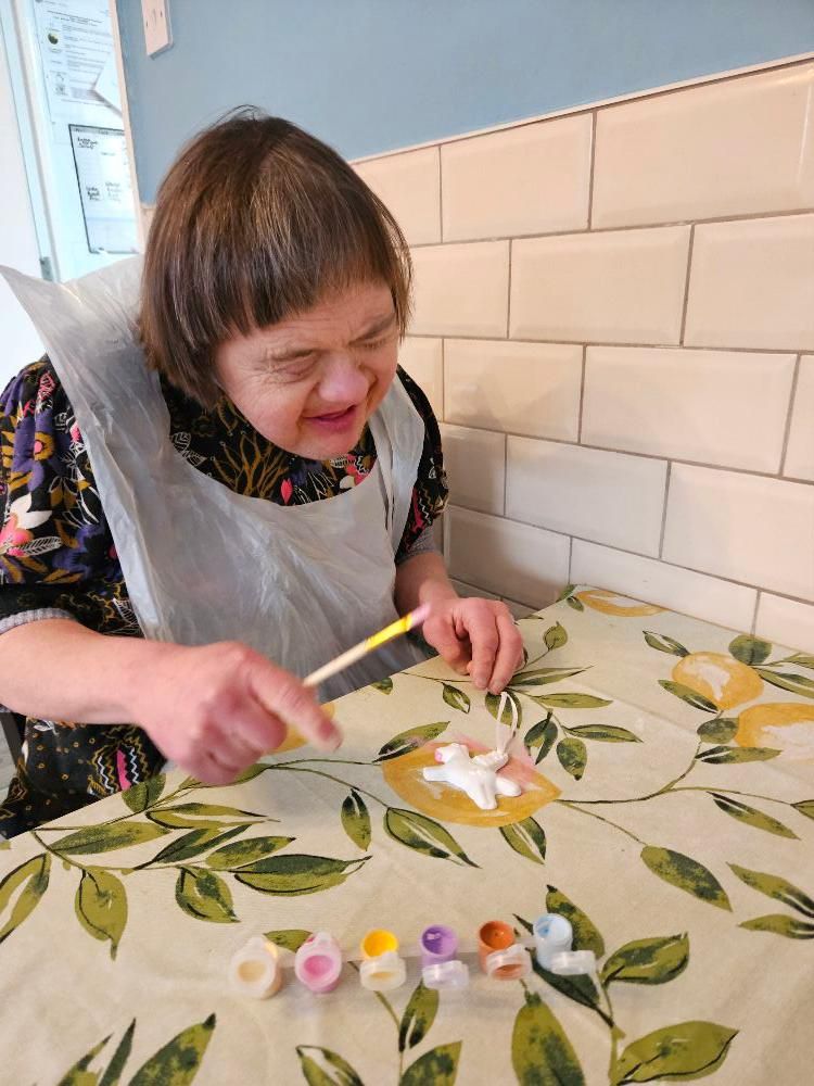 Woman with Down syndrome painting a figurine at a table, wearing a paint smock. Various colors of paint are visible.
