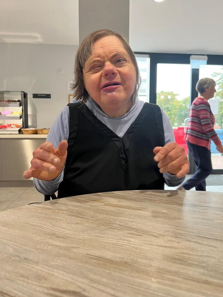 Woman with Down syndrome sitting, smiling at a table in a cafe, another person walking in the background.