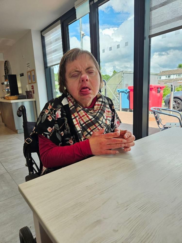 Woman in wheelchair at a table, making a face, wearing a floral shirt. Bright room with a window behind her.