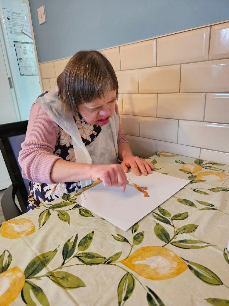 Person painting with a brush at a table with a lemon-patterned tablecloth. They wear a floral apron and pink shirt.