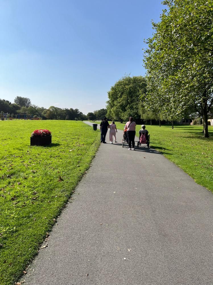 People walking on a paved path in a sunny park, one person using a wheelchair. Green grass, trees, blue sky.