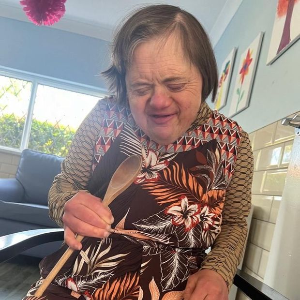 Woman with Down syndrome holding a wooden spoon in a kitchen, wearing a floral apron and smiling.