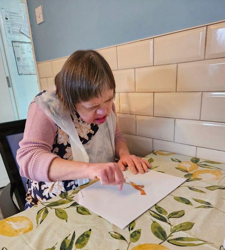Woman with Down syndrome painting at a table with floral tablecloth; kitchen setting.