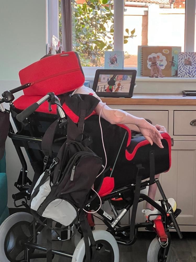 Person in a red and black wheelchair, resting arms on the sides. Tablet and books on a nearby surface.