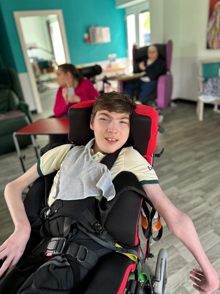 A young man in a wheelchair smiles; in a brightly lit room, two people sit at a table in the background.