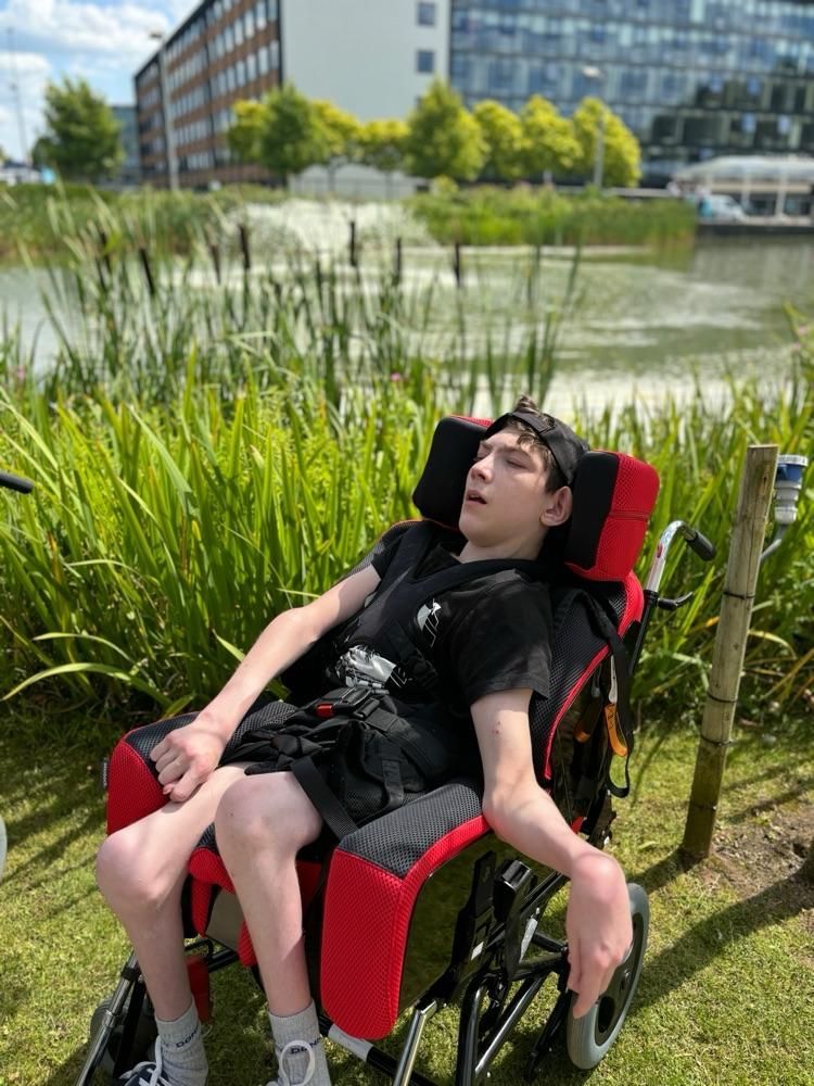 Person in a wheelchair near a pond with tall grass, looking up with mouth open. Buildings in background.