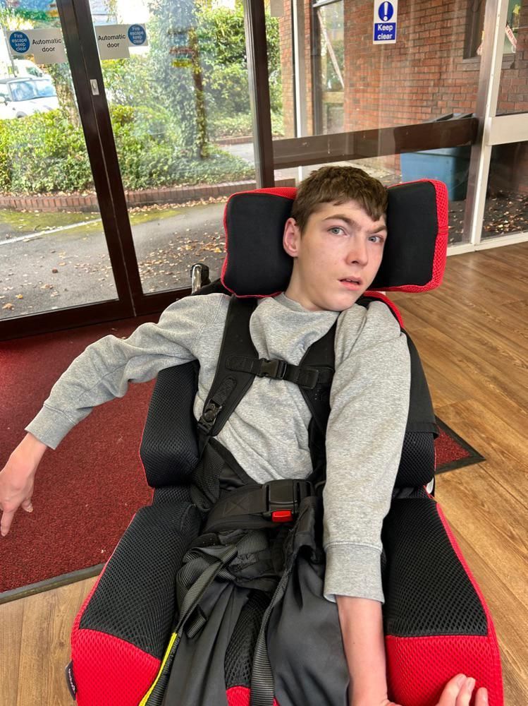 Young person in a wheelchair, wearing a grey shirt. The wheelchair has red accents, sitting near a glass door.