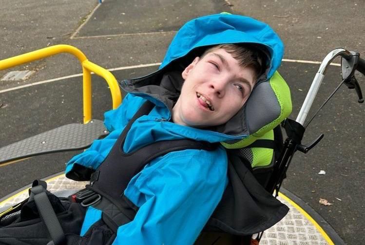 A young person in a wheelchair, wearing a blue hooded jacket, smiles on a playground carousel.