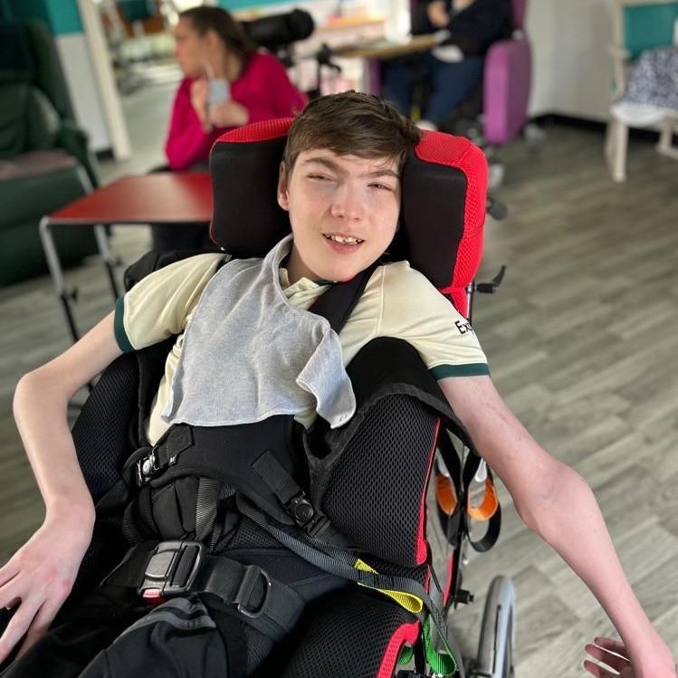 Young man in a wheelchair, smiling, wearing a gray top, in a care facility.