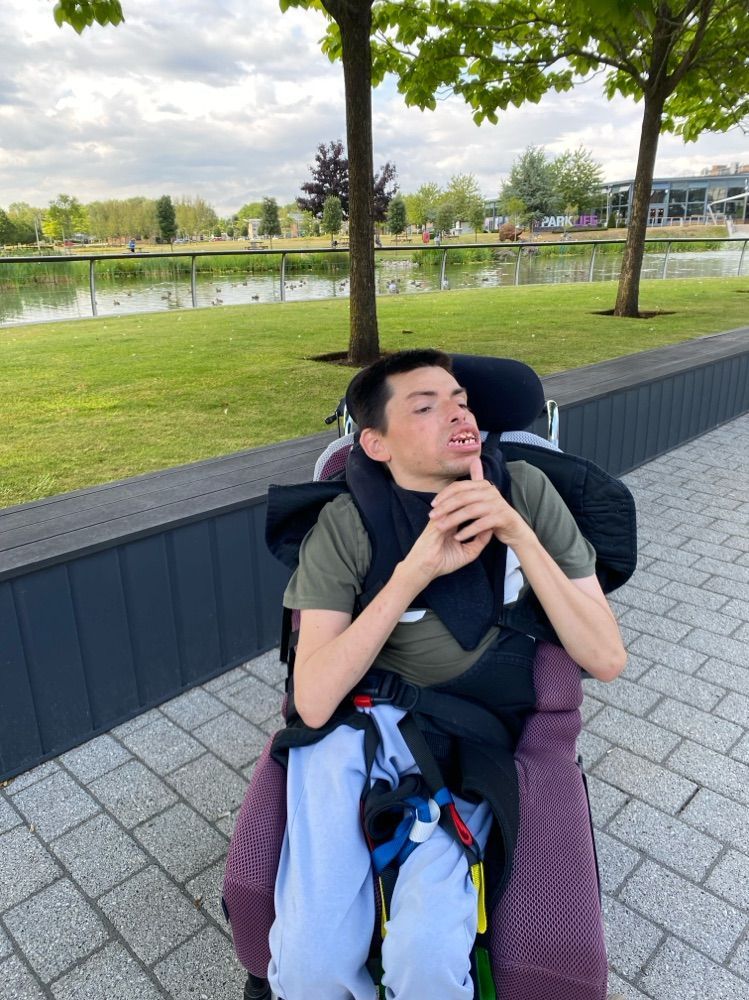 Man in wheelchair, olive green shirt, outside by a pond. He is looking up with an open mouth.