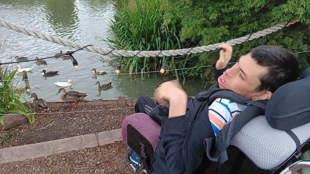 Person in wheelchair near pond with ducks, holding rope, smiling. Lush greenery and water in background.