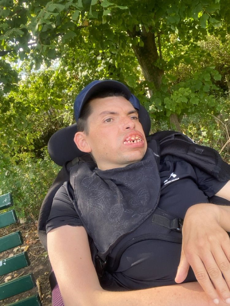 Man in a wheelchair wearing a blue cap and bib, outdoors near trees, smiling.
