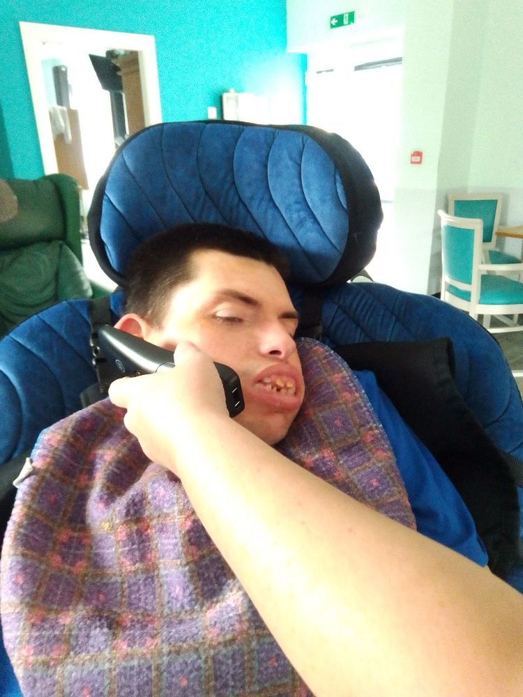 Person in wheelchair with bib, being assisted by a hand with a grooming tool; indoors.