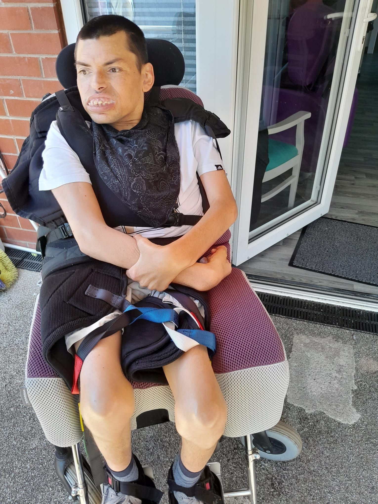 Man in wheelchair, looking to the left, sitting outside. Wearing a white shirt and black and purple coverings.