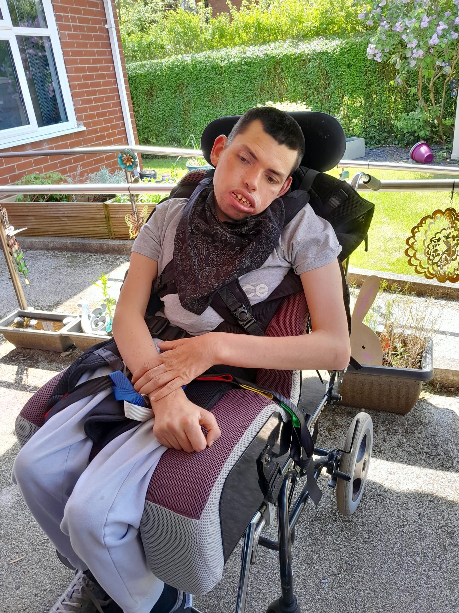 Person in a wheelchair, outdoors, wearing a scarf, looking at the camera, sunny day.