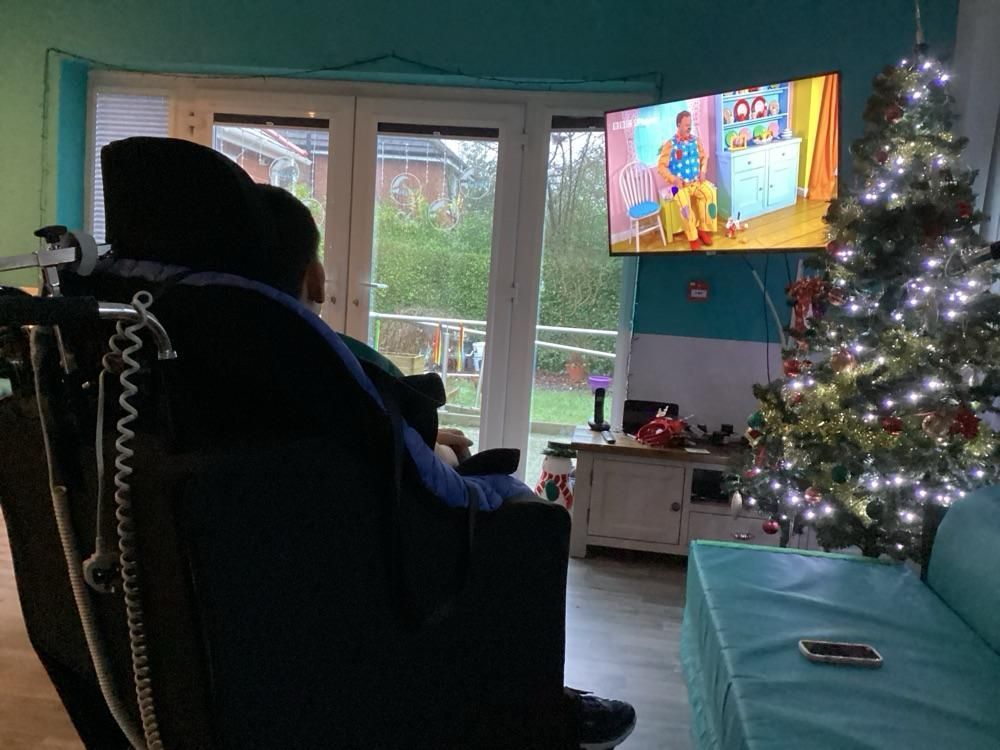 Person in wheelchair watches TV with a lit Christmas tree nearby in a living room with a blue and green color scheme.