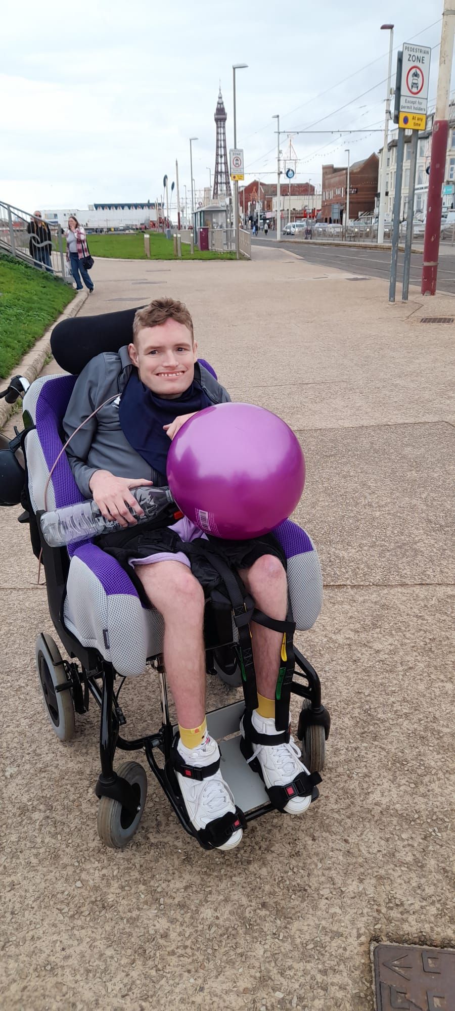 A young person in a wheelchair holds a purple balloon, smiling. They are on a paved walkway near a street.