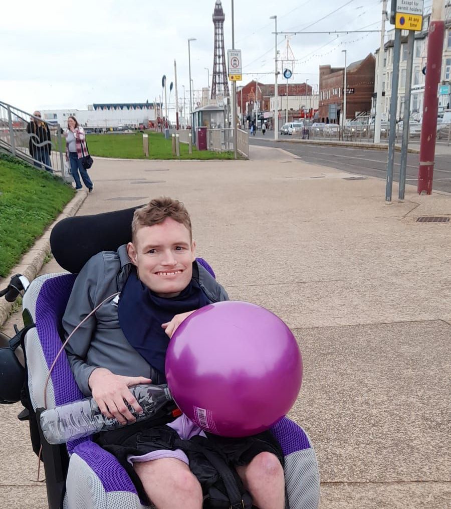 Man in wheelchair smiles, holds purple balloon, Blackpool Tower in background.