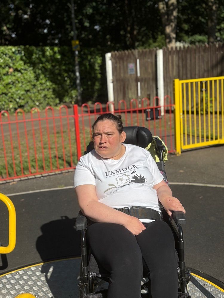 Woman in a wheelchair outside, wearing a white t-shirt. She is looking forward in a park setting with metal fences.