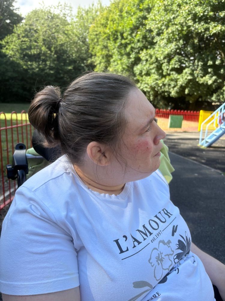 Woman in a white t-shirt with a ponytail, seated in a wheelchair, looks right, possibly at a playground.