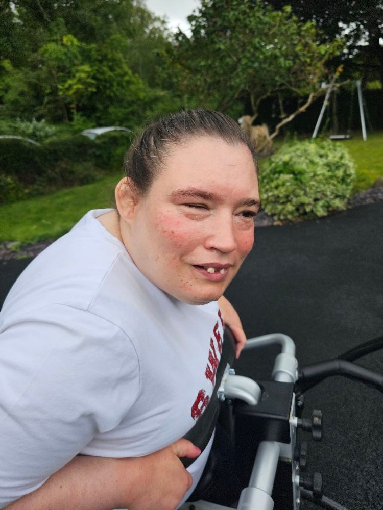 Woman in a wheelchair smiles outdoors, wearing a white shirt. Green trees and a playground are in the background.