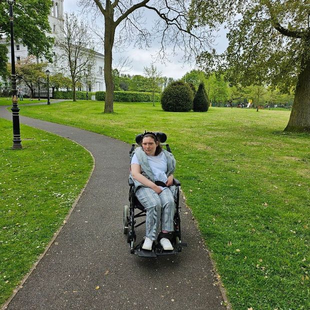 Person in a wheelchair on a path in a park. Green grass, trees, and a building are in the background.