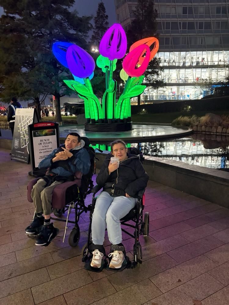 Two people in wheelchairs, in front of illuminated flower sculptures and a pond, at night.