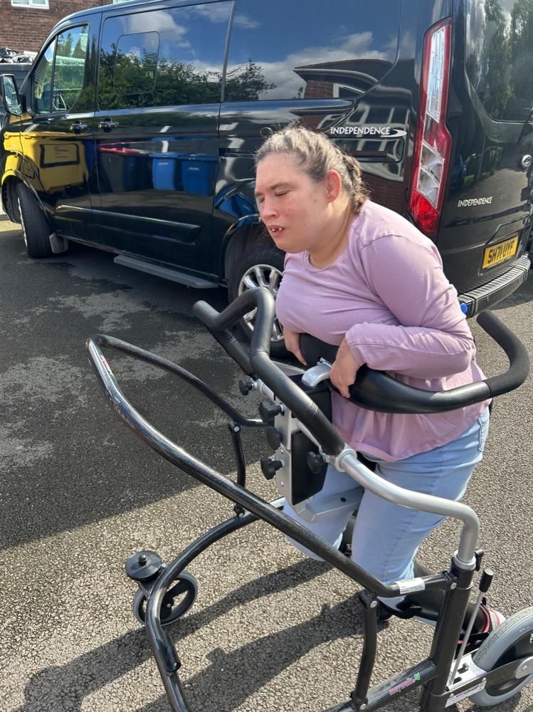 Woman with light skin using a walker in front of a black van. She wears a lavender shirt and jeans.