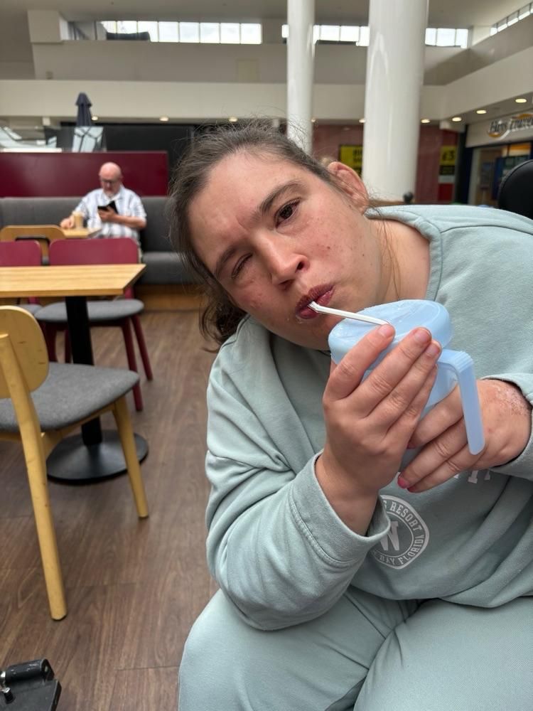 A person with a disability in a teal sweatshirt drinking from a sippy cup, seated. Restaurant interior.