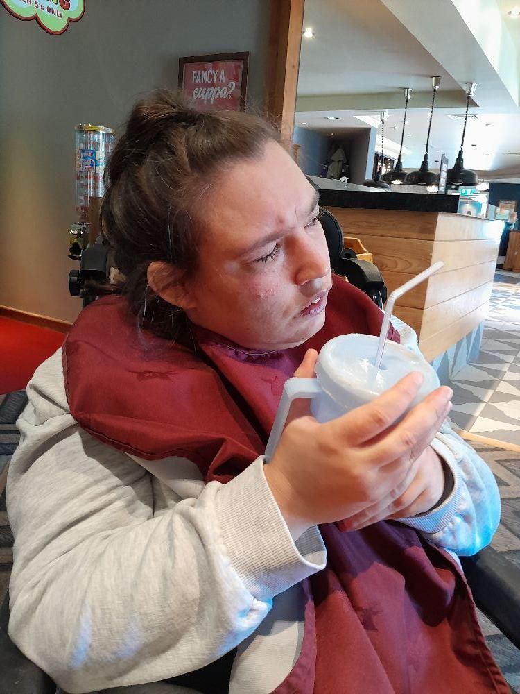Person in a wheelchair looking to the side, holding a cup with a straw, wearing a bib, indoors.