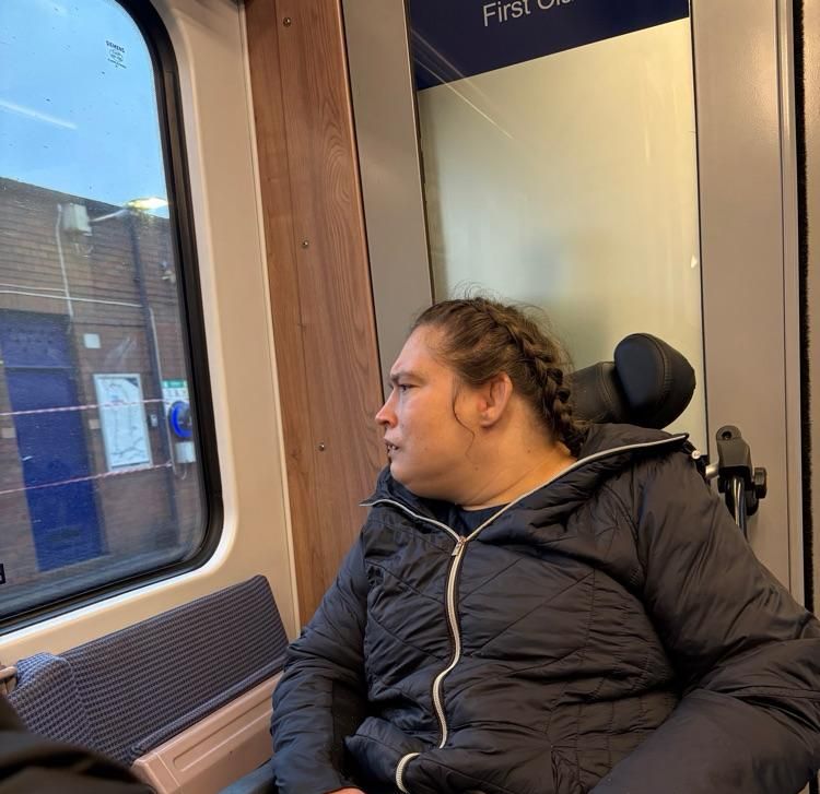 Woman in a wheelchair looking out a train window. Wearing a black coat, light skin, and braids.