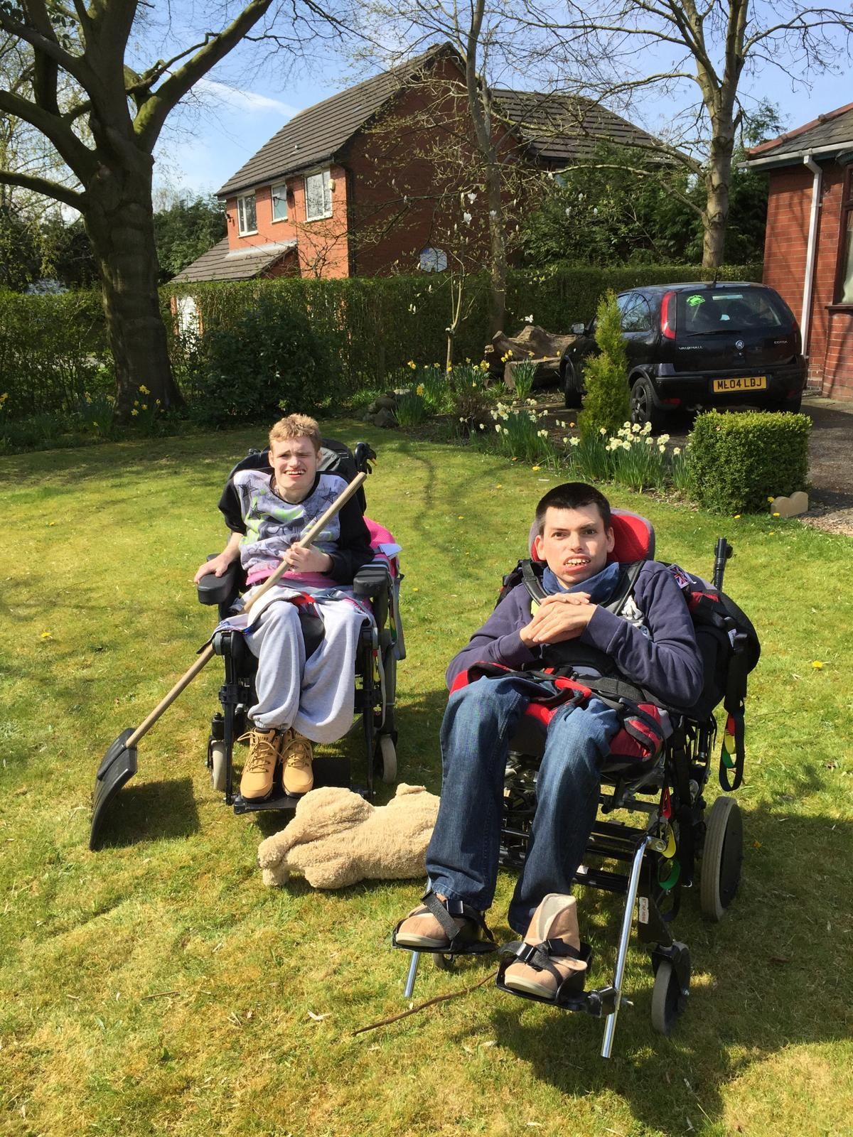 Two people in wheelchairs on a lawn, one holding a shovel, other with hands clasped. A house and car are in the background.