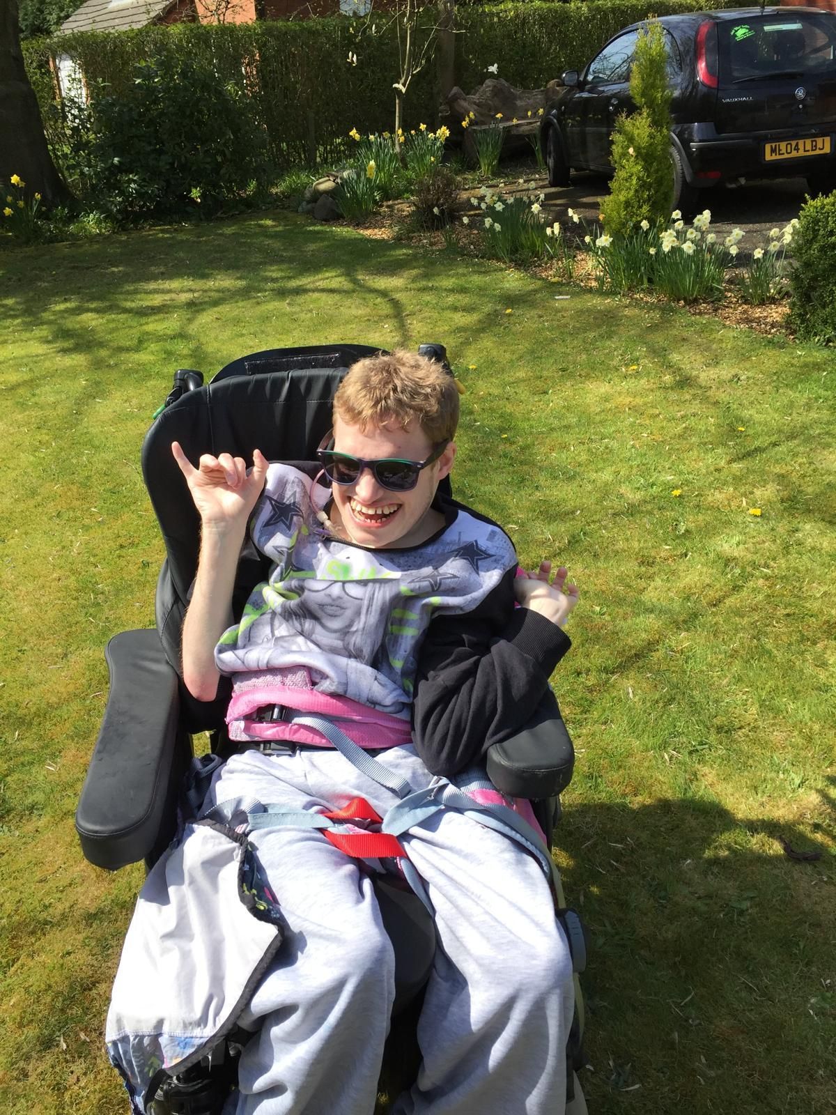 Person in a wheelchair smiling, wearing sunglasses, in a sunny garden.