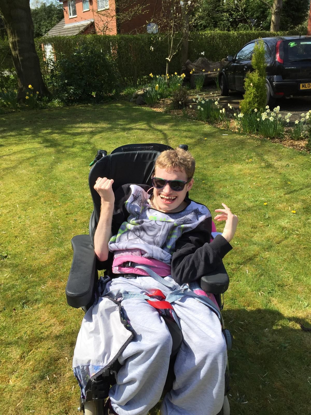 A young person in a wheelchair wearing sunglasses, smiling, in a sunny garden.