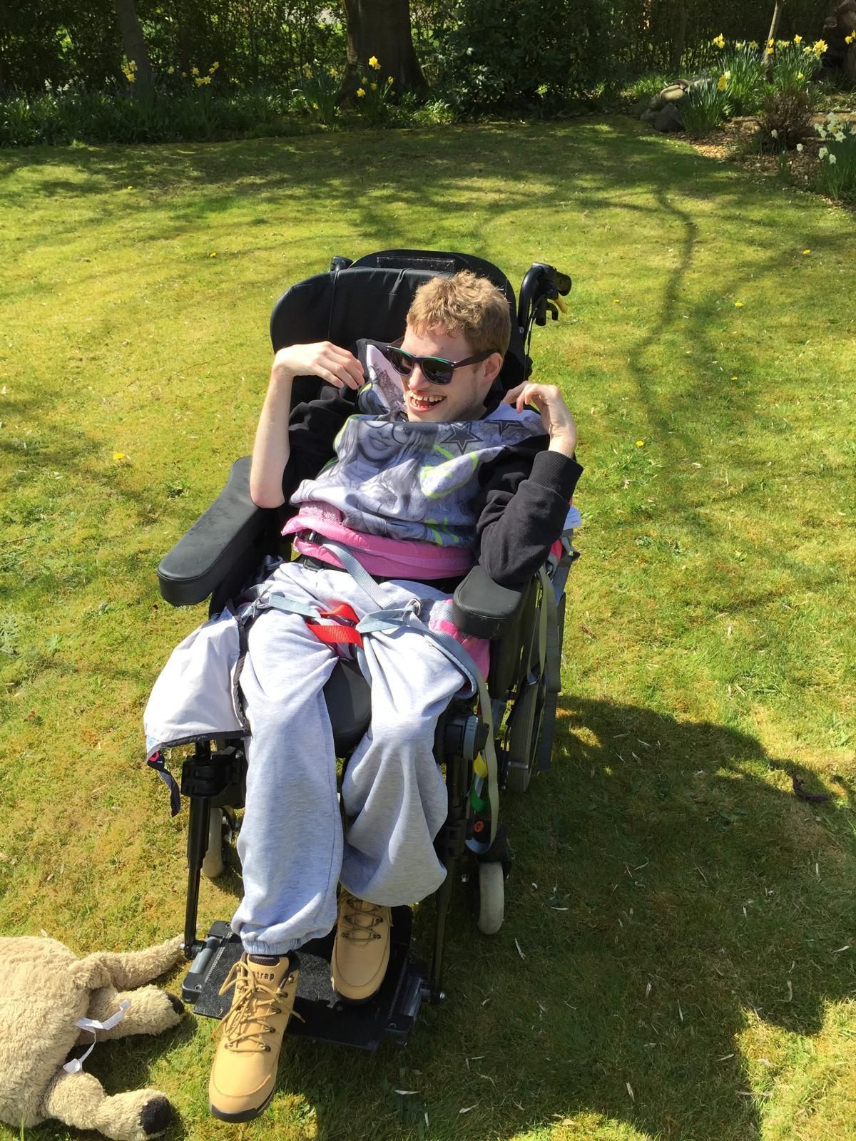 Young person in wheelchair outdoors, wearing sunglasses and smiling in the sunshine.