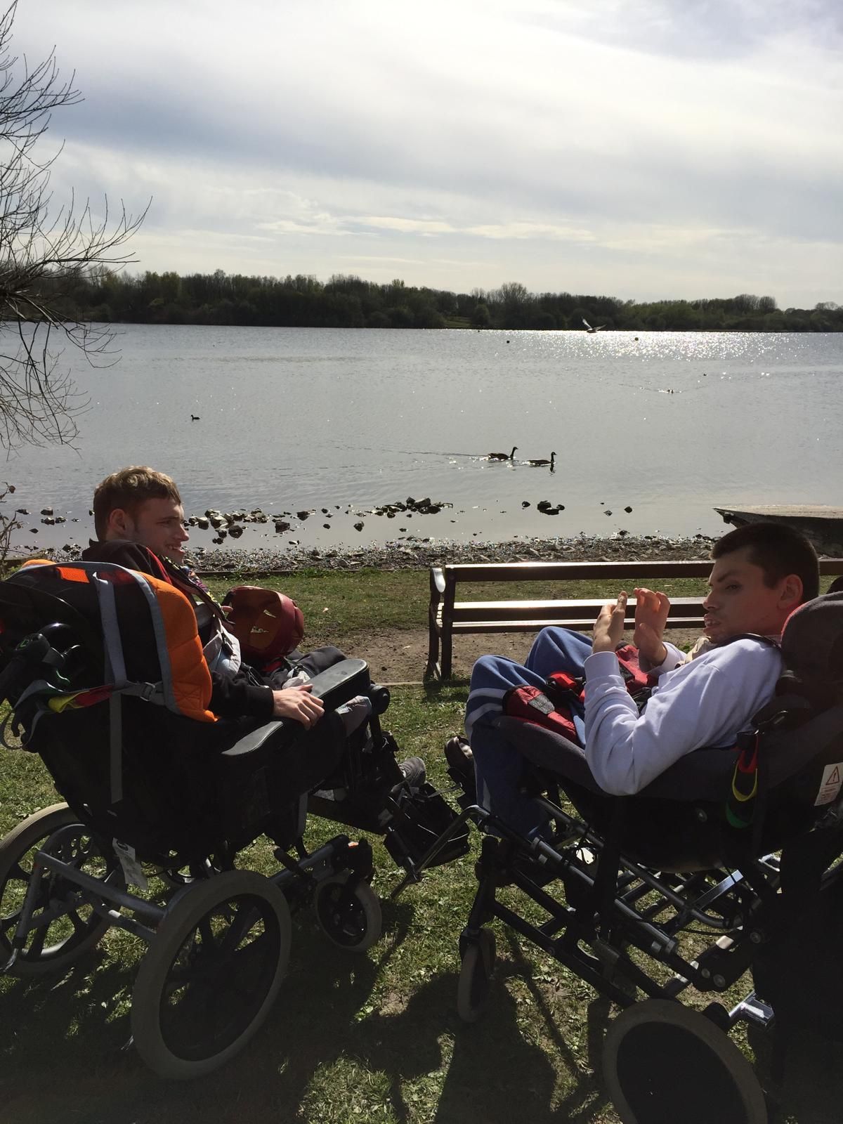 Two men in wheelchairs by a lake on a sunny day, conversing.