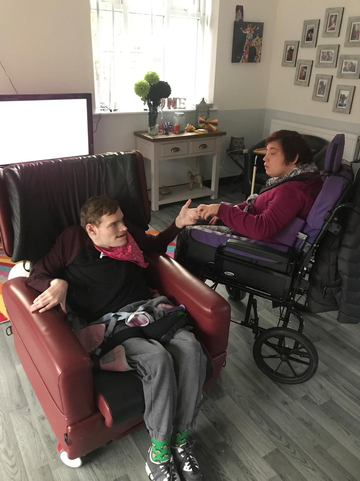 Two people, one in wheelchair, shake hands in living room. Red and purple clothing. Gray flooring.