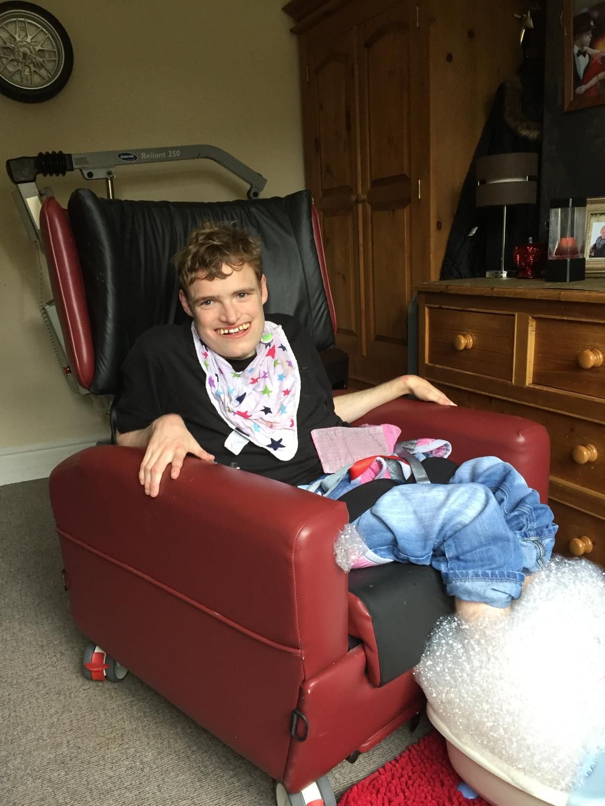 Person with disability smiling in a red chair, wearing a bib and jeans. Indoors, wooden furniture in background.