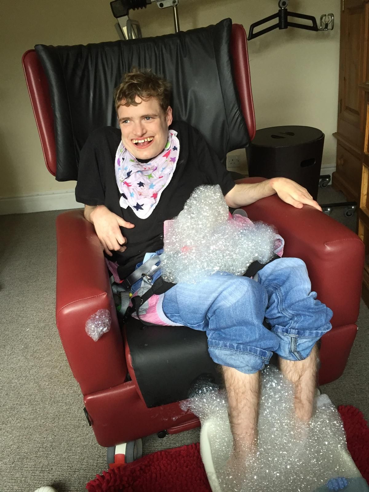 Person in a specialized red chair with bubbles. They smile and wear a bib, jeans, and black shirt. Indoor setting.