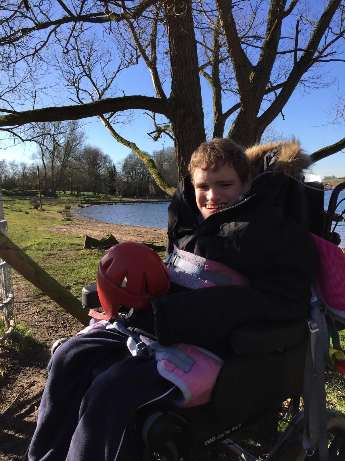 Person in a wheelchair smiles outside by a lake. They are wearing a black coat and holding a red helmet.