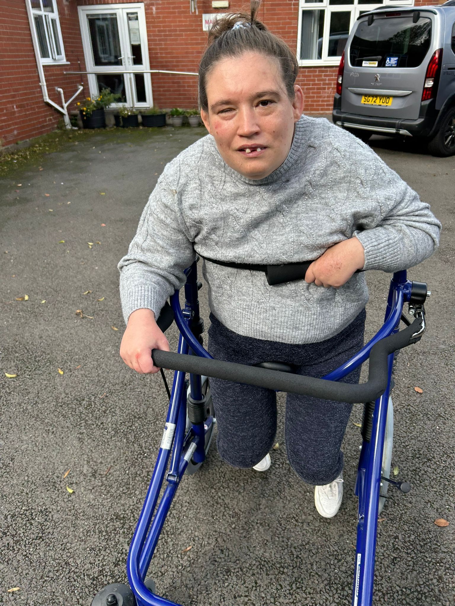 Woman with mobility aid, grey sweater, blue walker, and a van in the background.