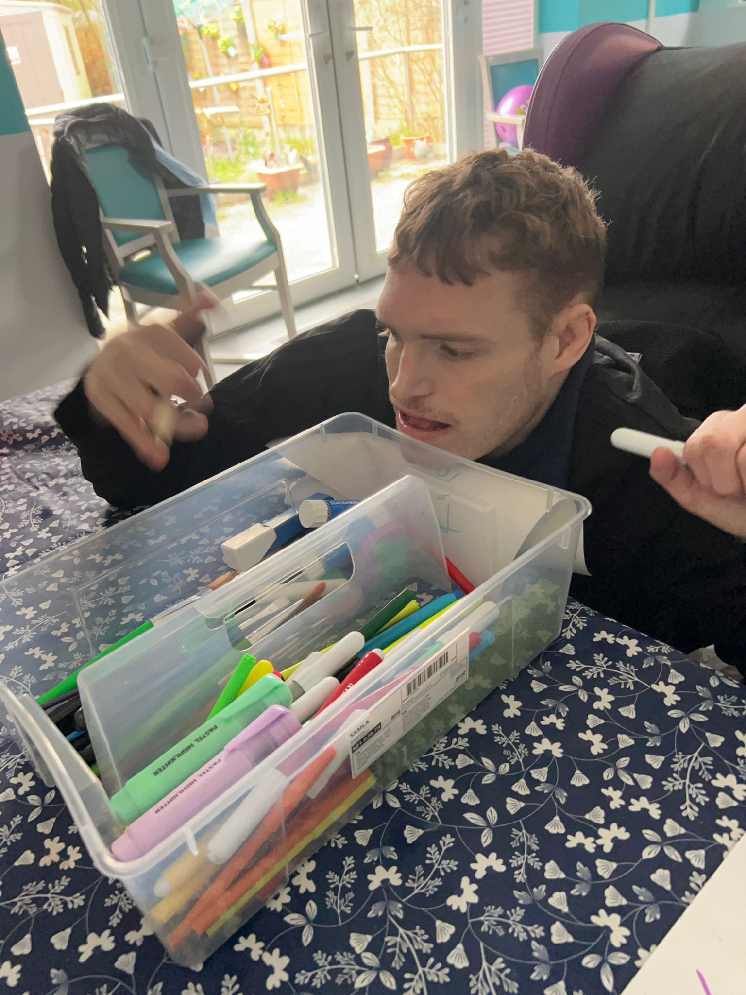 Young man lying down, looking at markers in a plastic bin. Indoor setting.
