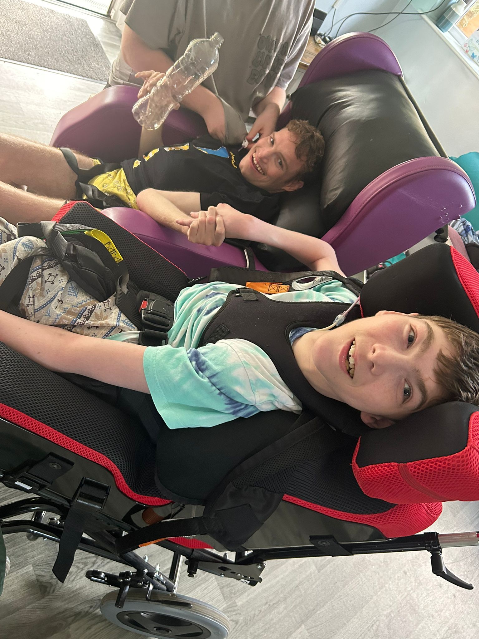 Two smiling boys, one in a wheelchair, holding hands, with an adult nearby. Purple cushions and gray floor.