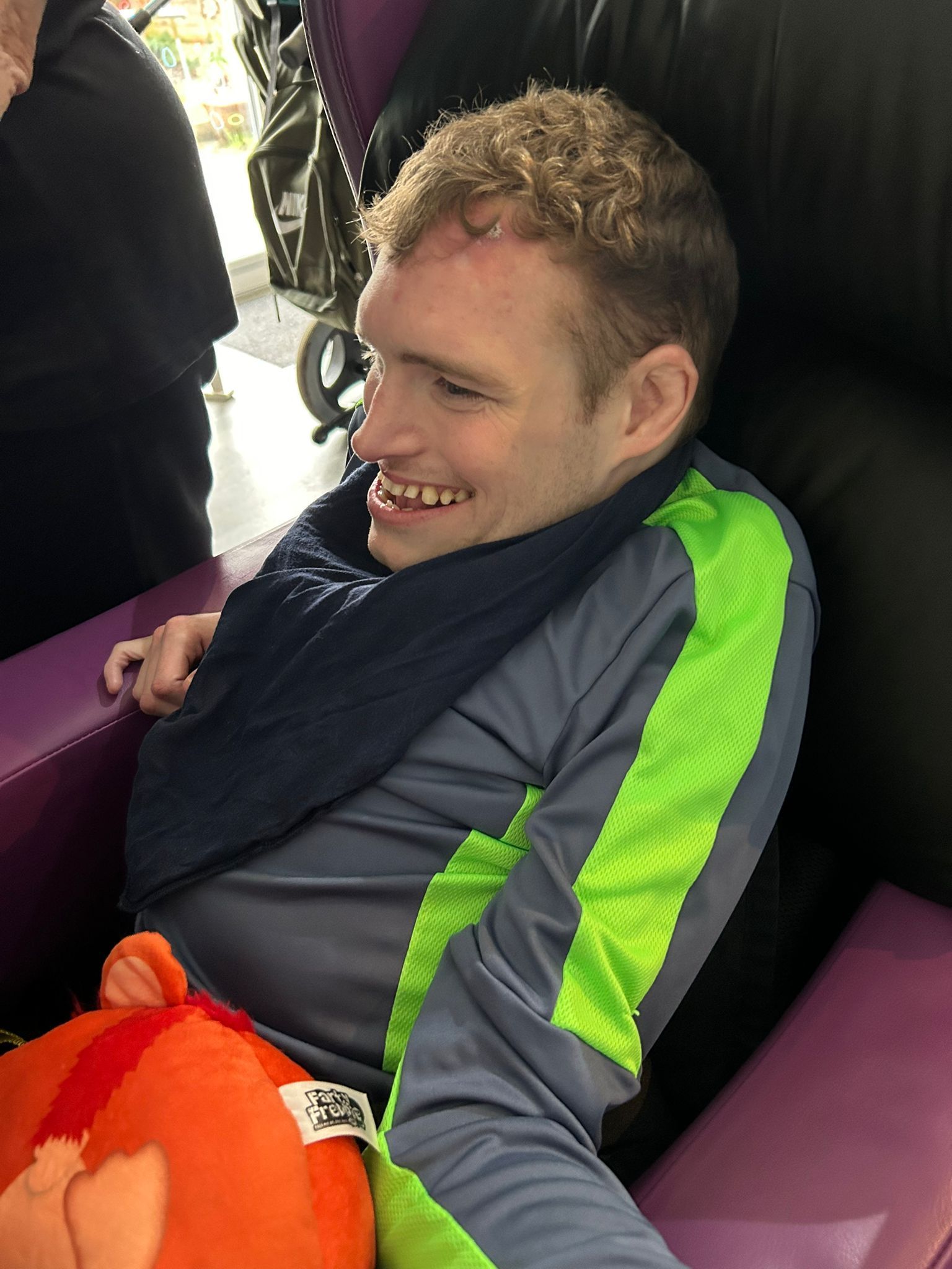 Man with curly hair smiling, sitting in a chair, wearing a gray shirt, a dark scarf, and holding an orange plushie.