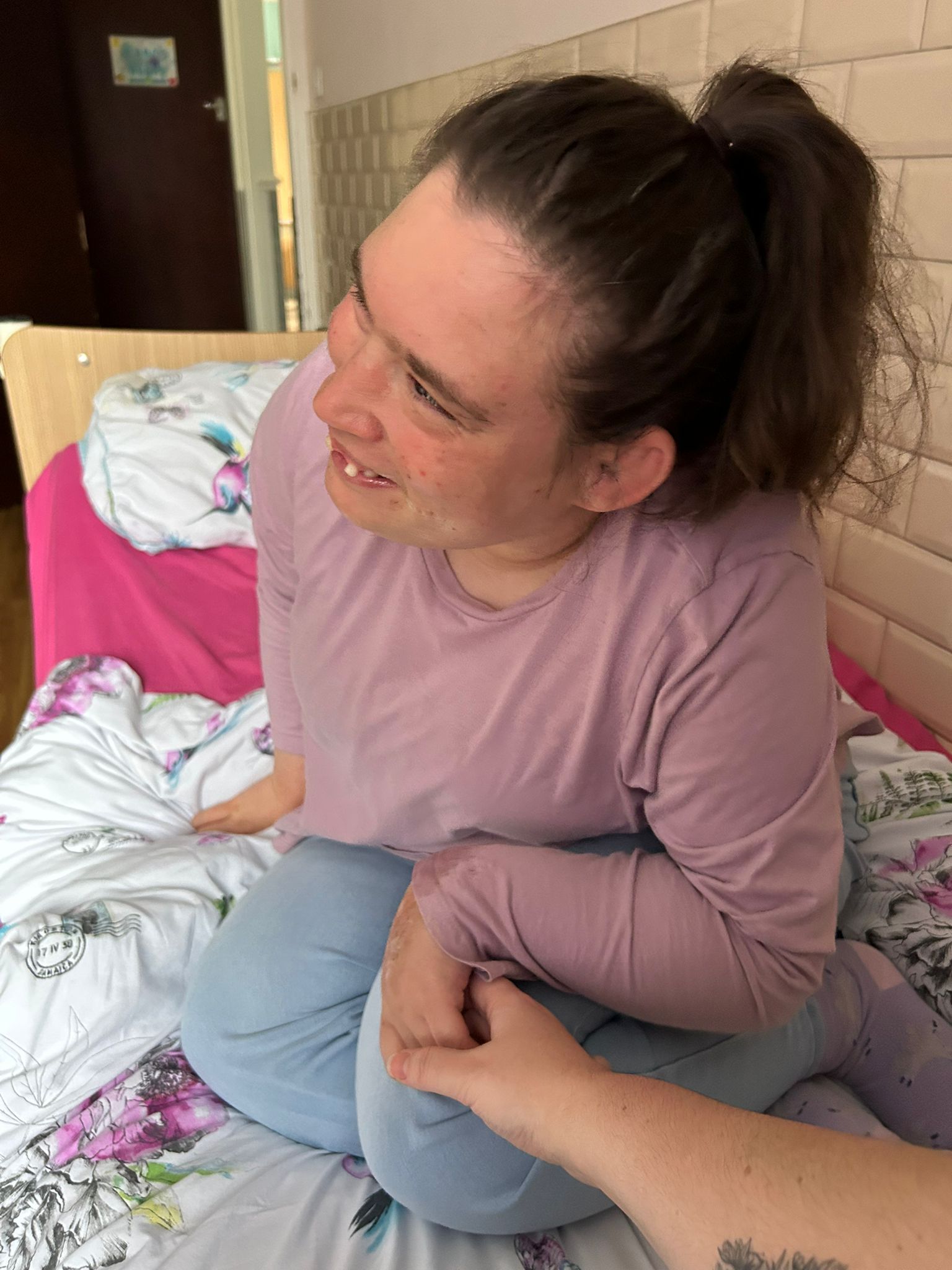 Woman in pink top and jeans, smiling, sitting on floral bed, looking at a hand.