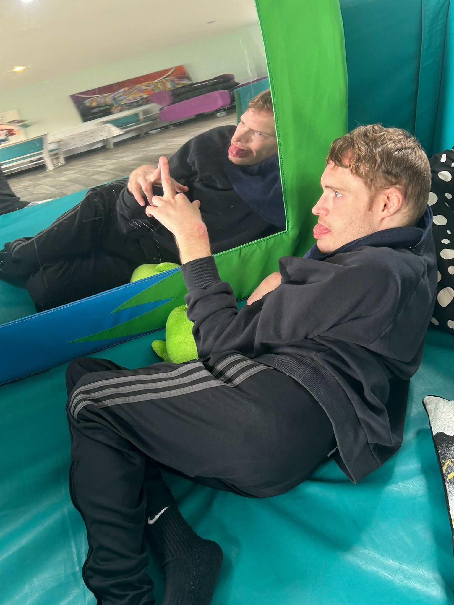 Man in black sweatsuit looking at himself in a mirror. He is indoors and has a playful expression.
