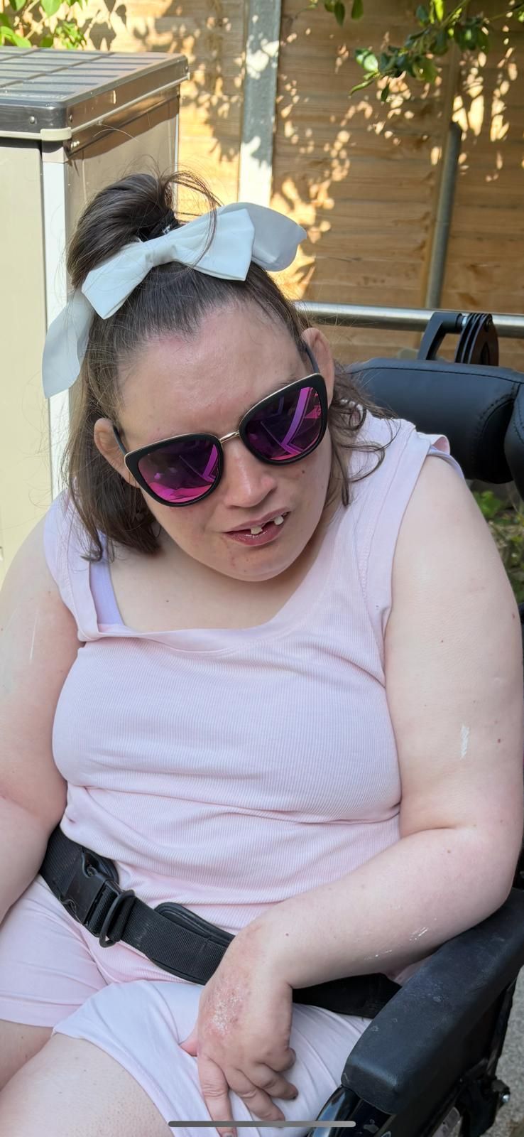Woman in sunglasses, wheelchair, pink dress, and bow in her hair, outside.